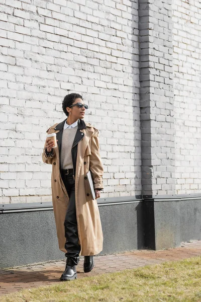 Full length of african american businesswoman in sunglasses and trench coat walking with laptop and coffee to go near grey wall on urban street — Stock Photo