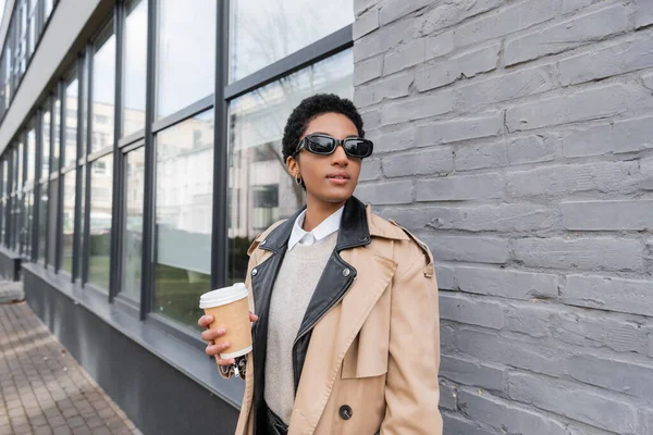 Africano americano empresária em óculos de sol e casaco bege trincheira segurando takeaway bebida e olhando para longe perto de edifício na rua da cidade — Fotografia de Stock