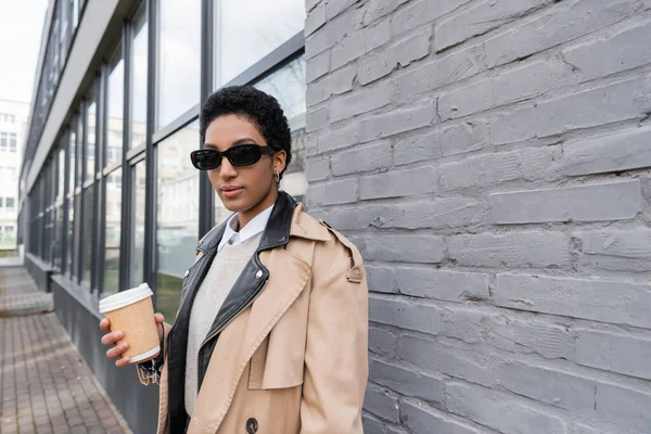 Afrikanisch-amerikanische Geschäftsfrau in Sonnenbrille und beigem Trenchcoat steht mit Kaffee in der Nähe einer grauen Hauswand auf der Straße — Stockfoto