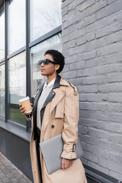 African american businesswoman in stylish trench coat and sunglasses standing with coffee to go and laptop near grey wall of city building — Stock Photo