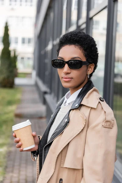 Young african american businesswoman in  sunglasses and trench coat holding coffee to go near blurred building on street — Stock Photo