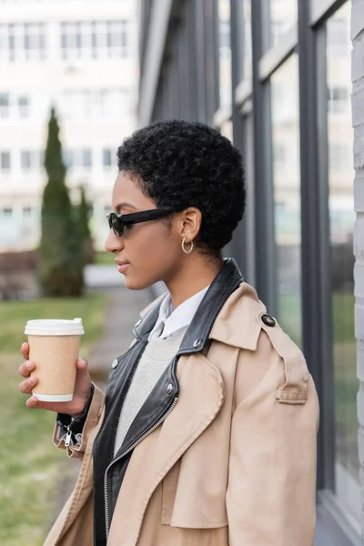 Seitenansicht einer afrikanisch-amerikanischen Geschäftsfrau in Sonnenbrille und beigem Trenchcoat, die mit Imbiss auf der Straße steht — Stockfoto