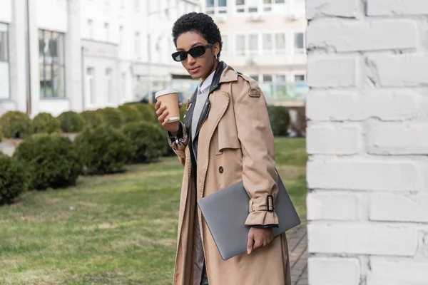 Mulher de negócios americana africana na moda com bebida takeaway e laptop olhando para a câmera perto do edifício — Fotografia de Stock