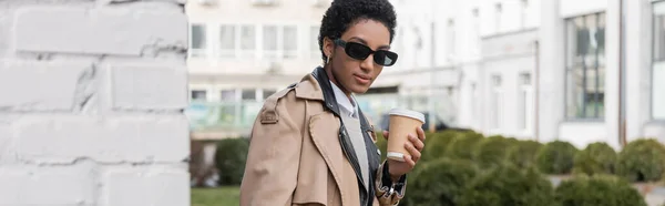 Élégante femme d'affaires afro-américaine en lunettes de soleil et trench coat tenant tasse en papier et regardant la caméra sur la rue de la ville, bannière — Photo de stock