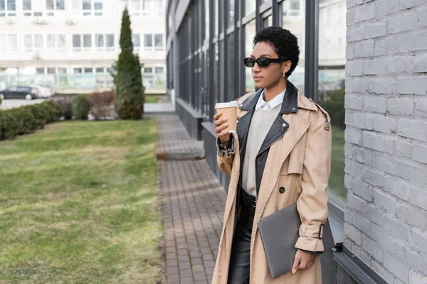 Mujer de negocios afroamericana de moda en gafas de sol de pie con portátil y bebida para llevar cerca del edificio de la ciudad con fachada de vidrio — Stock Photo