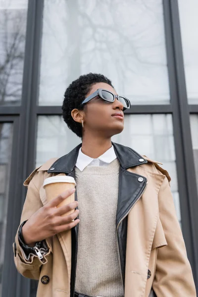 Low angle view of african american businesswoman in trendy sunglasses holding coffee to go near city building with glass facade — Stock Photo
