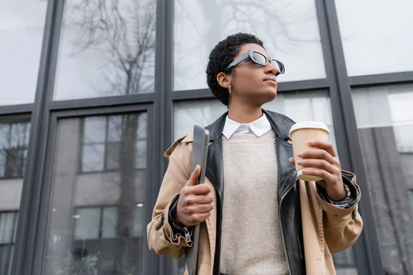 Visão de baixo ângulo da mulher de negócios americana africana na moda com laptop e bebida takeaway olhando para longe perto da fachada de vidro na rua urbana — Fotografia de Stock