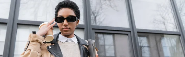 Joven mujer de negocios afroamericana con portátil y taza desechable ajustando gafas de sol de moda cerca del edificio de la ciudad con fachada de vidrio, pancarta - foto de stock