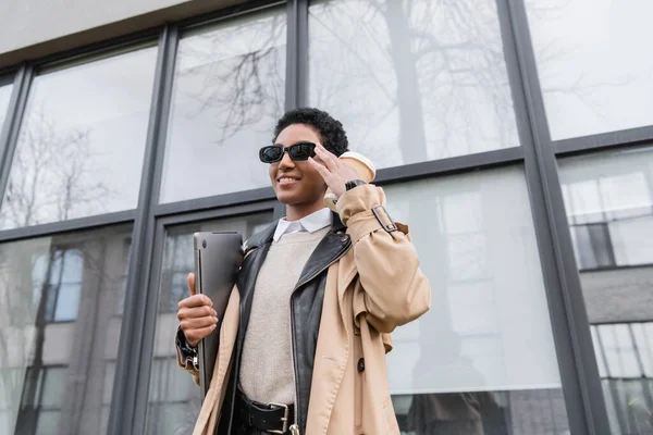 Visão de baixo ângulo de feliz mulher de negócios afro-americana em óculos de sol de pé com copo de papel e laptop perto de edifício urbano com fachada de vidro — Fotografia de Stock