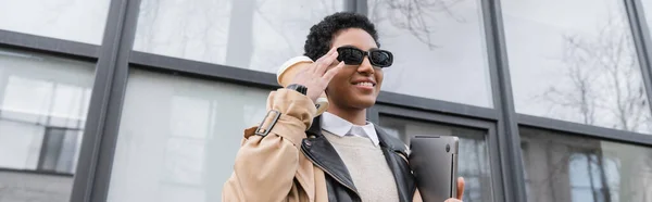 Alegre mujer de negocios afroamericana con café para llevar y portátil ajuste de gafas de sol cerca del edificio, pancarta - foto de stock