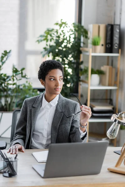 Thoughtful african american businesswoman in suit holding eyeglasses while sitting at workplace in office — Stock Photo