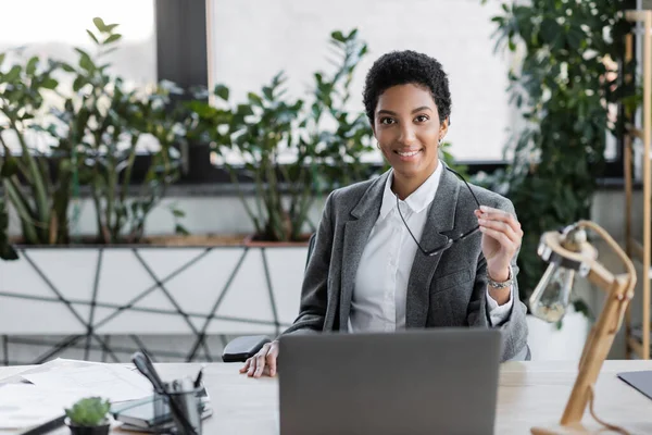 Радісна афроамериканська бізнесменка в сірому костюмі, яка тримає окуляри і дивиться на камеру біля ноутбука на робочому столі в офісі. — стокове фото
