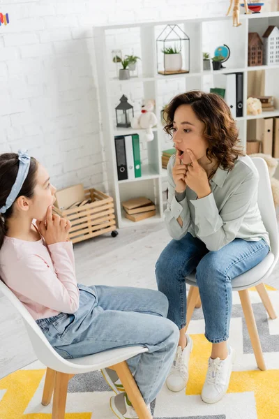 Terapeuta del habla hablando y tocando mejillas durante la lección con el niño en la sala de consulta - foto de stock
