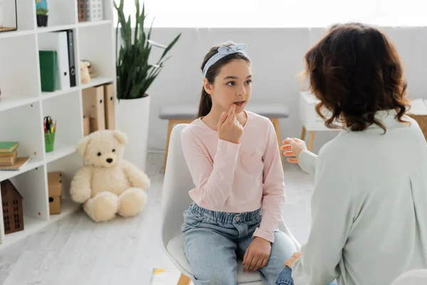 Preteen girl talking and pointing at mouth near blurred speech therapist in consulting room — Stock Photo
