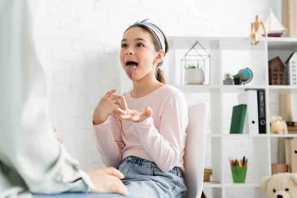 Ученик-подросток, торчащий языком рядом с размытым логопедом в кабинете консультанта — стоковое фото