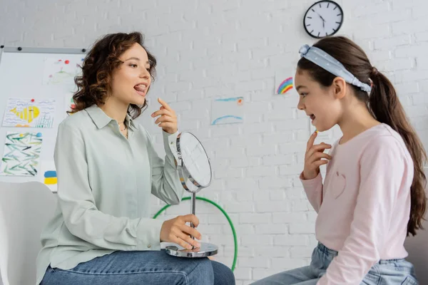 Speech therapist holding mirror and pointing at mouth near pupil in consulting room — Stock Photo