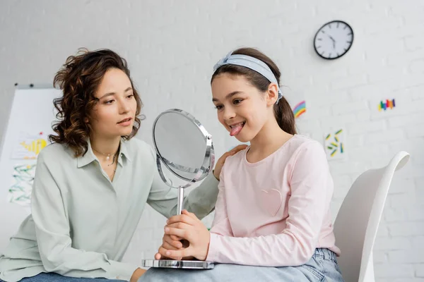 Speech therapist looking at pupil sticking out tongue and holding mirror in consulting room — Stock Photo