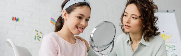 Девочка-подросток, торчащая языком возле зеркала и логопеда в кабинете консультанта, баннер — стоковое фото