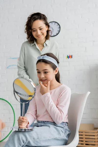 Preteen child holding mirror and speaking near smiling speech therapist in consulting room — Stock Photo