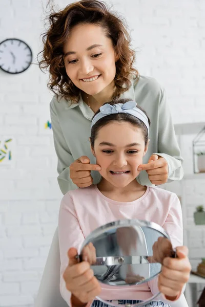 Smiling speech therapist touching cheeks of preteen child with mirror in consulting room — Stock Photo