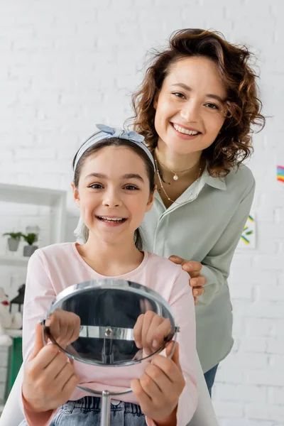 Позитивный ребенок держит зеркало рядом с логопедом в кабинете консультанта — стоковое фото