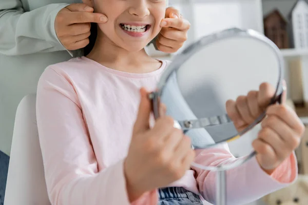 Cropped view of speech therapist touching cheeks of smiling pupil with mirror in consulting room — Stock Photo