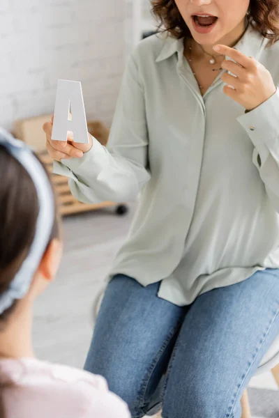 Cropped view of speech therapist holding letter a and pointing at mouth near blurred pupil in consulting room — Stock Photo