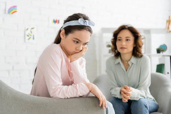 Upset preteen girl sitting near blurred psychologist in consulting room — Stock Photo
