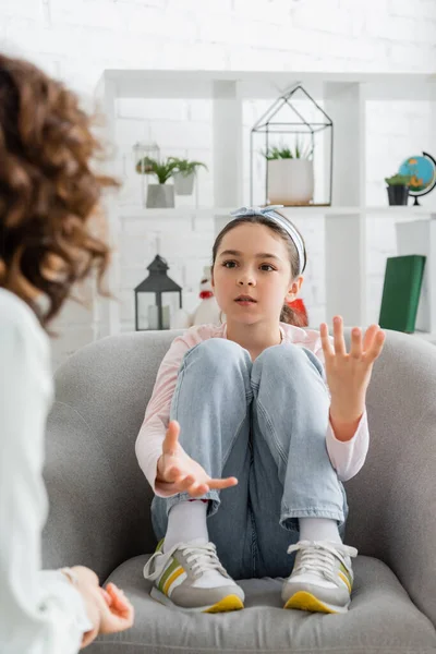 Chica preadolescente hablando con psicólogo borroso mientras está sentado en el sillón en la sala de consulta - foto de stock