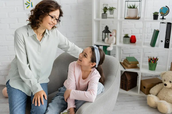 Cheerful psychologist in eyeglasses looking at happy preteen patient in consulting room — Stock Photo