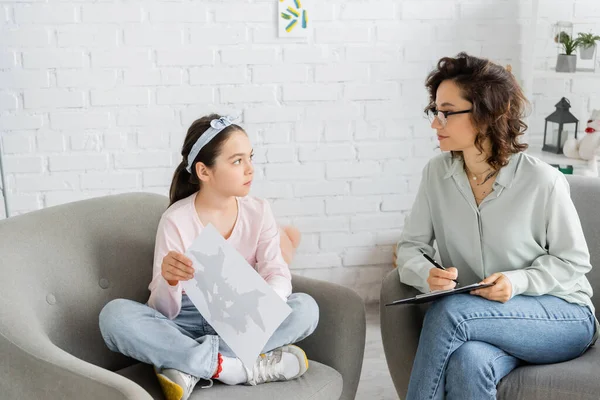 Preteen girl holding Rorschach test near psychologist with clipboard in consulting room — Stock Photo