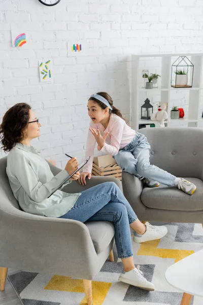 Garota pré-adolescente excitada conversando com psicólogo sorridente com área de transferência no consultório — Fotografia de Stock