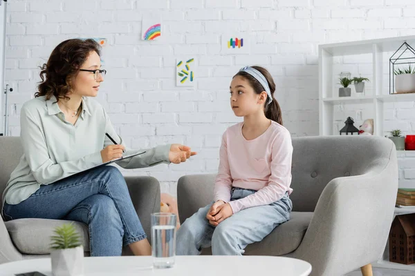 Psicólogo escribiendo en portapapeles y hablando con niña preadolescente en la sala de consulta - foto de stock