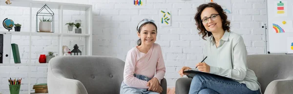Cheerful psychologist with clipboard and preteen child looking at camera in consulting room, banner — Stock Photo