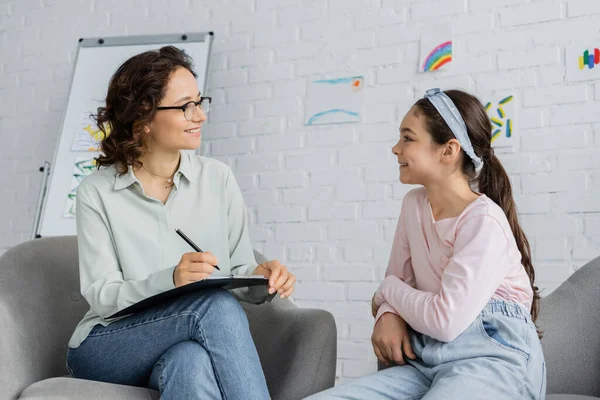 Smiling psychologist in eyeglasses holding clipboard and looking at preteen daughter in consulting room — Stock Photo