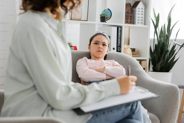 Angry girl looking at blurred psychologist with clipboard in consulting room — Stock Photo