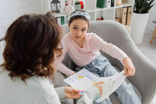Preteen patient talking to blurred psychologist pointing at drawing in consulting room — Stock Photo