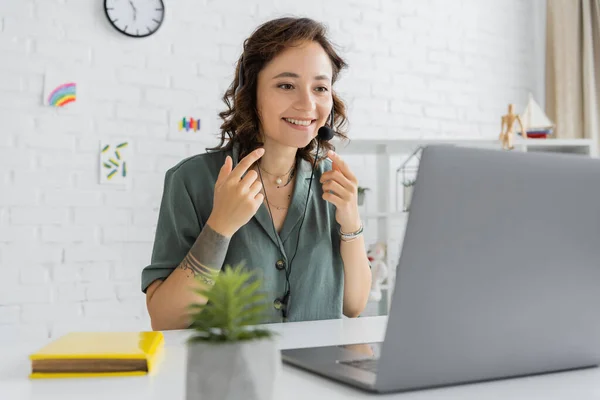 Улыбающийся логопед в наушниках, указывающий пальцами во время видеозвонка на ноутбук в консультационной комнате — стоковое фото