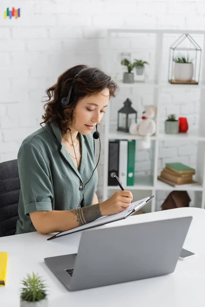 Speech therapist in headset writing on clipboard near laptop in consulting room — Stock Photo