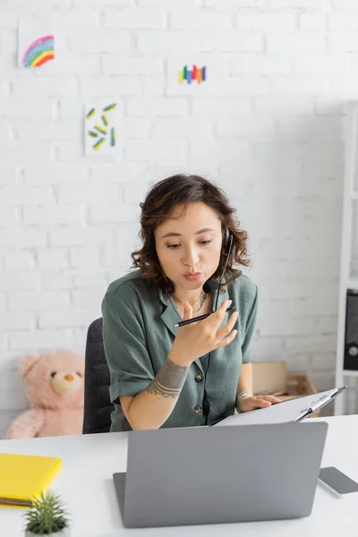 Speech therapist in headset holding clipboard and talking during video call in consulting room — Stock Photo
