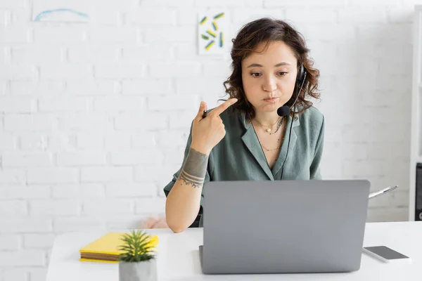Speech therapist in headset pointing at mouth and holding clipboard during video call on laptop in consulting room — Stock Photo