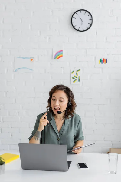 Speech therapist in headset talking and holding clipboard during video call on laptop in consulting room — Stock Photo