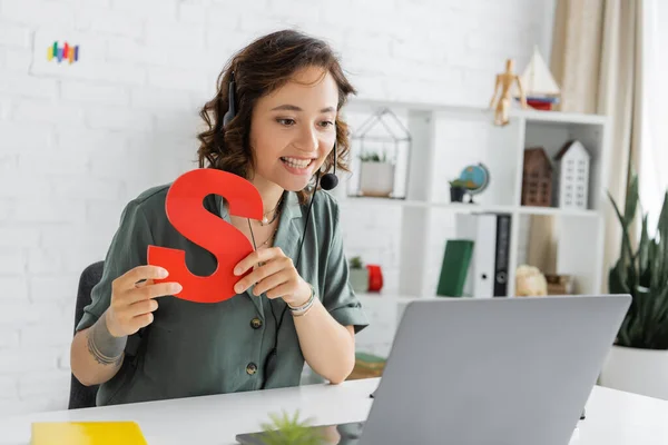 Smiling speech therapist in headset holding letter s during video call on laptop in consulting room — Stock Photo