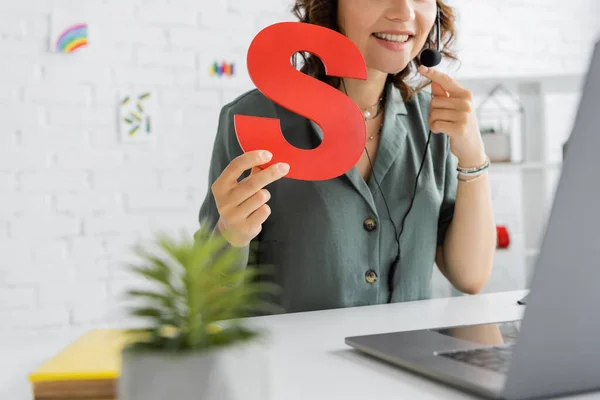 Cropped view of smiling speech therapist in headset holding letter s during video call on laptop — Stock Photo