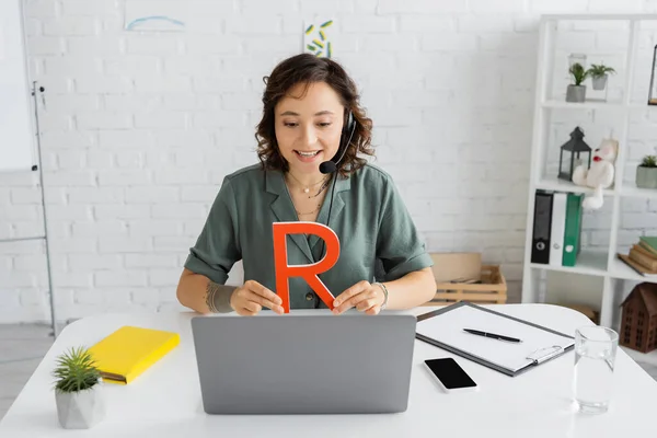 Positive Logopädin im Headset hält Brief neben Laptop während Videoanruf im Sprechzimmer — Stockfoto