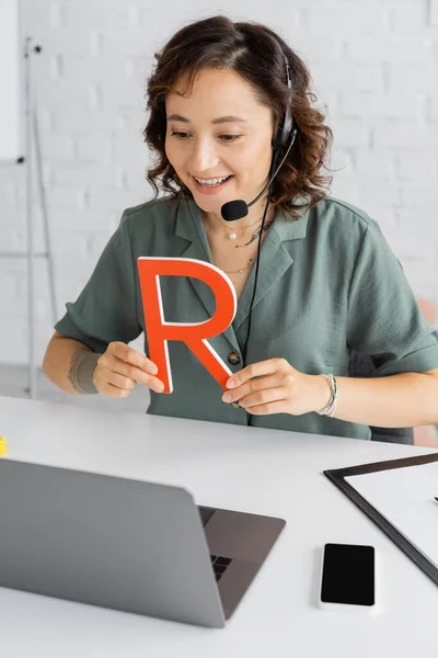 Cheerful speech therapist in headset holding letter during video lesson on laptop in consulting room — Stock Photo