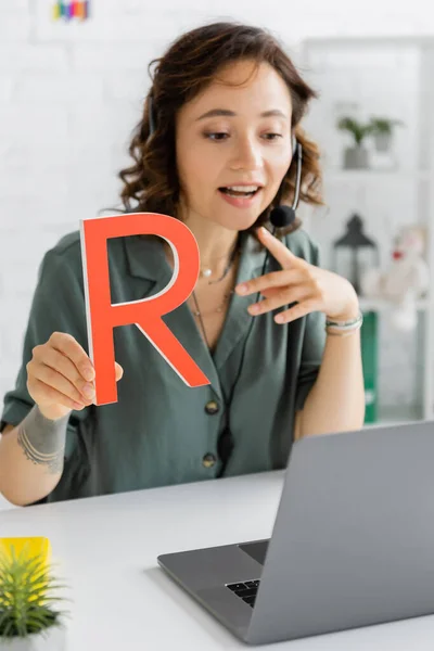 Blurred speech therapist in headset holding letter during online lesson in consulting room — Stock Photo