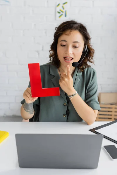 Speech therapist in headset pointing at tongue and holding letter during online lesson on laptop — Stock Photo