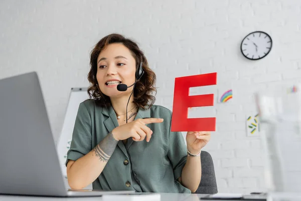 Smiling speech therapist in headset pointing at letter during online lesson on laptop in consulting room — Stock Photo