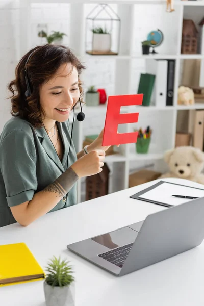 Overjoyed speech therapist in headset holding letter during video call on laptop in consulting room — Stock Photo